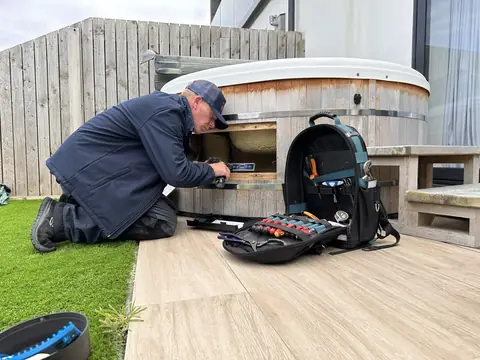 Jon Pennington kneeling at a cedar hot tub with the access panel open and his tool bag beside him